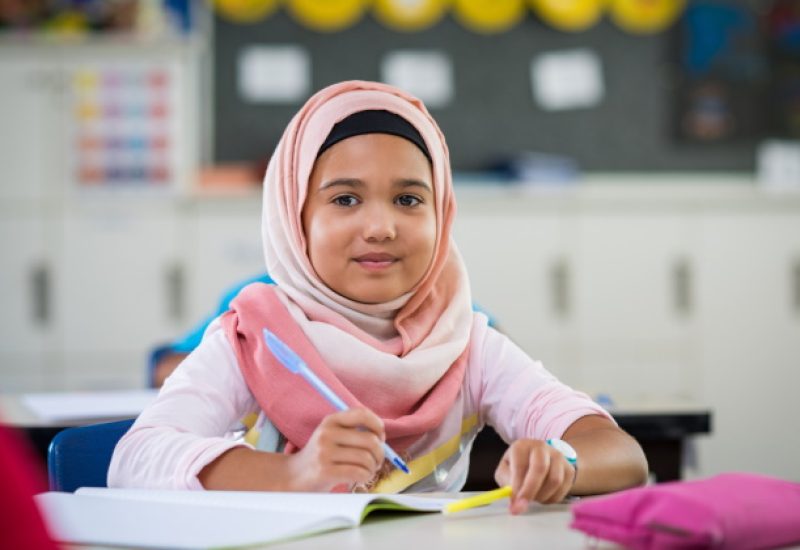 Happy young smiling girl wearing hijab and looking at camera while sitting at desk with book and pen in hand. Elementary muslim schoolgirl writing notes in classroom. Portrait of arab school girl in chador. (Happy young smiling girl wearing hijab and