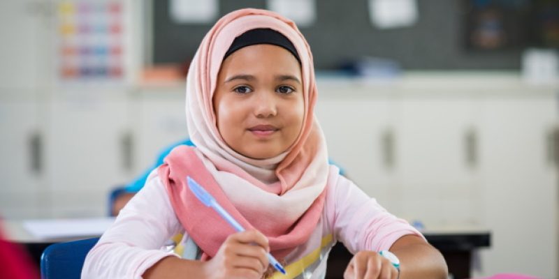 Happy young smiling girl wearing hijab and looking at camera while sitting at desk with book and pen in hand. Elementary muslim schoolgirl writing notes in classroom. Portrait of arab school girl in chador. (Happy young smiling girl wearing hijab and