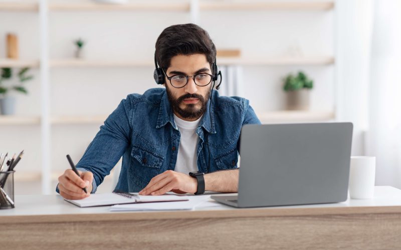 Millennial arab guy in headset watching online webinar on laptop at home, having online training, studying with computer and taking notes to notepad, copy space